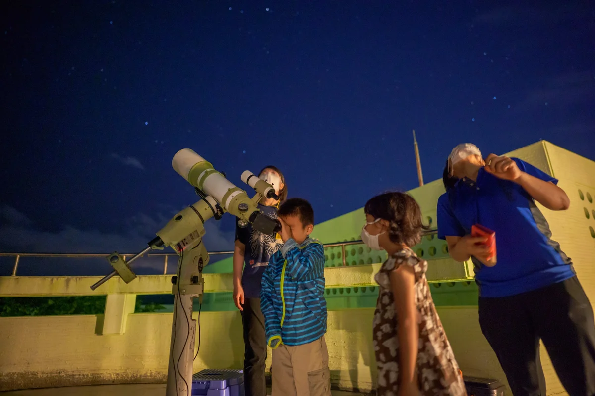 星空の下、子どもが望遠鏡を覗き、周りの人たちが空を見上げている